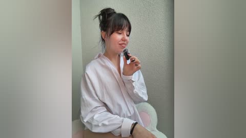 A video of a young Asian woman with light skin, wearing a white robe, sitting against a textured wall, holding a black pen, with a serene expression, in a minimalist room.