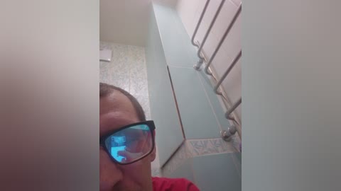 A video captures a person in a bathroom, wearing blue-tinted glasses and a red shirt, with a mirror reflecting a white tiled wall and a metallic towel rack.