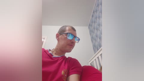 Video of a bald man with blue glasses, wearing a red t-shirt and gold necklace, sitting on a red couch in a room with patterned wallpaper.