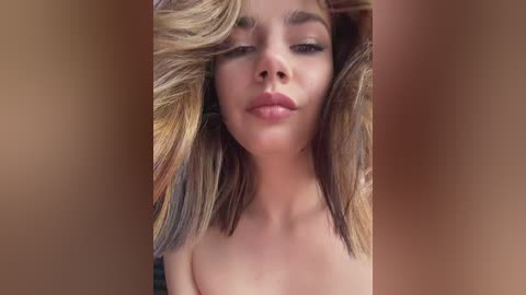 Video of a young, fair-skinned woman with shoulder-length, blonde hair dyed with multicolored streaks. She has a neutral expression, wearing no makeup. The background is a blurred, warm brown, focusing attention on her face.