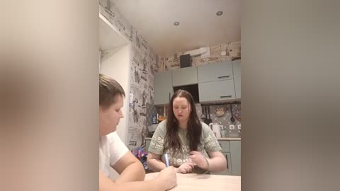 Video of a woman with long brown hair in a green sweater, seated at a light wood table in a rustic kitchen with patterned wallpaper and grey cabinets.