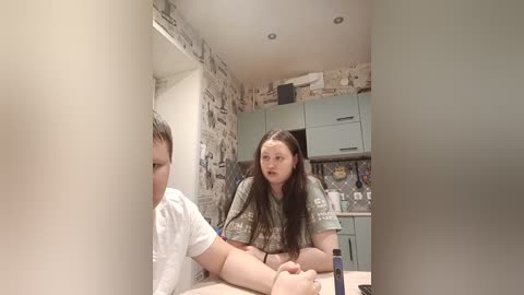 Video of a young Caucasian woman with long dark hair, wearing a camo shirt, sitting at a kitchen counter. A young boy in a white shirt stands beside her. The kitchen has a rustic, cluttered ambiance with pastel-colored cabinets and a stone-patterned backsplash.