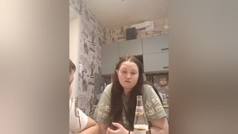 Video of a woman with long black hair, wearing a green t-shirt, sitting in a room with newspaper-covered walls and a kitchen background. She holds a glass of beer.