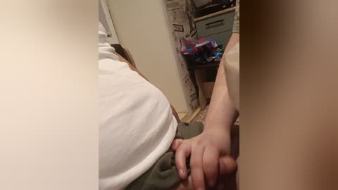 Video of a person's white t-shirt, with a hand holding a smartphone, partially visible. Background shows a cluttered kitchen with a cluttered countertop and open cabinet.