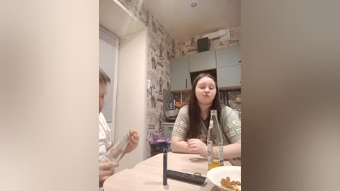 Video of a young woman with long brown hair, seated at a table in a modern kitchen with light gray cabinets, eating a sandwich.