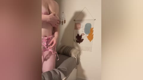 A video of a pale-skinned woman in pink lingerie, standing against a beige wall with a hanging art piece featuring abstract leaves.
