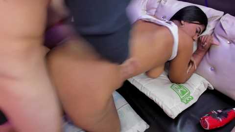 Video of a nude woman with light skin and dark hair, lying on a bed, wearing only a white tank top. A blurred male figure is partially visible in the foreground.