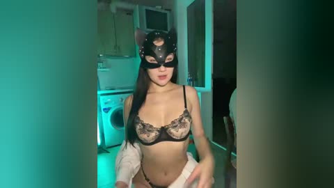 Video of an East Asian woman with long black hair, wearing a black cat mask and lace lingerie, sitting in a dimly lit kitchen with green lighting.