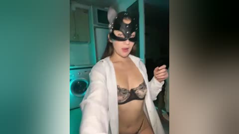 A video of a woman in a cat mask, white lab coat, and lacy black lingerie, standing in a dimly lit kitchen with a washing machine in the background.