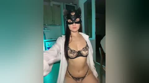 Video of a light-skinned woman in black lace lingerie, black mask, and white robe, standing in a dimly lit kitchen with teal lighting.