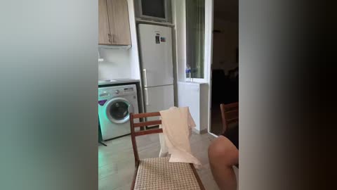 Video of a modern, minimalist kitchen with a wooden chair, white towel draped over it, and a white fridge. The setting is bright, clean, and sparsely furnished.