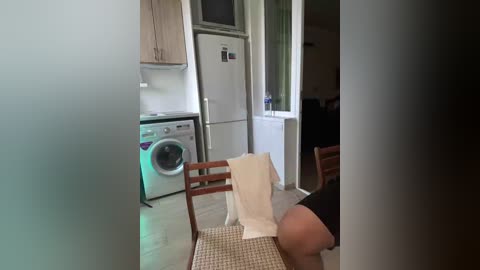 A video of a small, modern kitchen with white appliances, wooden cabinets, and a single wooden chair with a white towel draped over it. The scene is partially obscured by a frosted glass door.