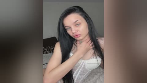 Video of a young Caucasian woman with long black hair, fair skin, and a slender physique, wearing a white lace corset, standing in a modern bedroom with neutral-colored walls and a bed.