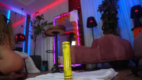 Video of a dimly-lit, cozy room with a yellow can, a pink couch, plants, and a drum set under colorful, multicolored LED lights.