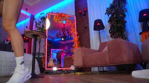 Video of a modern, dimly lit living room with purple and blue LED lighting, featuring a person in white socks and sneakers, a plush armchair, and a vibrant floral arrangement in the background.