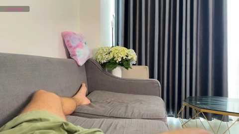 A video of a light-skinned person in green shorts, lounging on a gray sofa with a floral pillow, against black and white striped curtains, a vase of white hydrangeas, and a modern glass-topped coffee table.