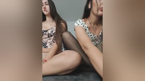 A video of two young women with fair skin, one in a polka-dot top and the other in a leopard print top, sitting close on a grey couch.