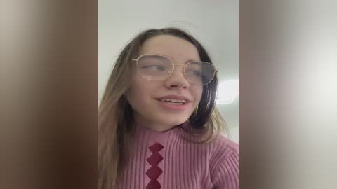 Video of a young Caucasian woman with straight, shoulder-length brown hair, wearing wire-rimmed glasses and a pink ribbed sweater with diamond-shaped red accents. She smiles slightly, with a neutral expression.