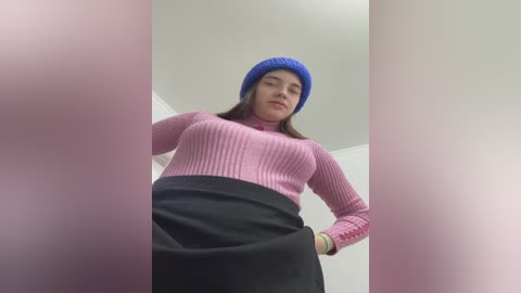Video of a young woman with medium skin tone, wearing a blue knit beanie, pink ribbed sweater, and black skirt, hands on hips, indoors, low angle shot.