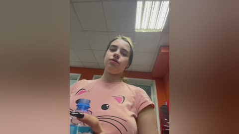 Video of a young woman with light skin and blonde hair, wearing a pink t-shirt with a cartoon cat face, drinking from a blue water bottle. Background includes orange walls and fluorescent lights.