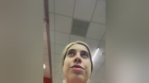 Video of a blonde woman with fair skin, blue eyes, and pouty lips, looking upward in a brightly lit office with white ceiling tiles and fluorescent lights.