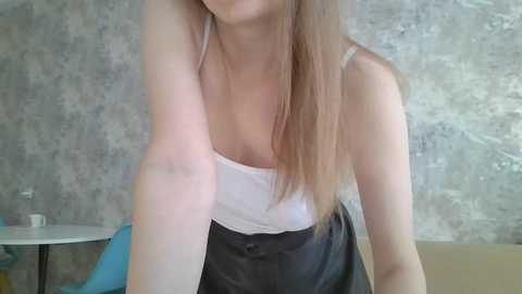 Video of a Caucasian woman with light skin and long, straight brown hair, wearing a white tank top and black skirt, posing indoors with a textured gray wall background.