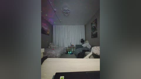 Video of a dimly lit, sparsely furnished bedroom with two beds, one with a green lamp, white curtains, and a star-shaped ceiling light.