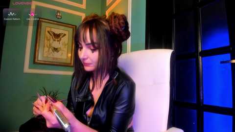 A video of a young woman with dark hair in a bun, wearing a black leather jacket, seated in a white chair. She holds a small plant in her hands. The background features teal walls and framed artwork.