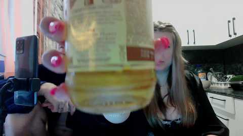 A blurry video of a young woman with long, blonde hair in a black robe, holding a beer bottle in front of her face, taken in a modern kitchen with white cabinets and dark countertops.