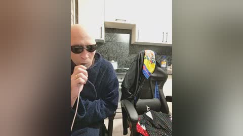 Video of an elderly man in dark glasses, wearing a blue robe, sitting in a black gaming chair in a modern kitchen. A colorful hat is on the chair.