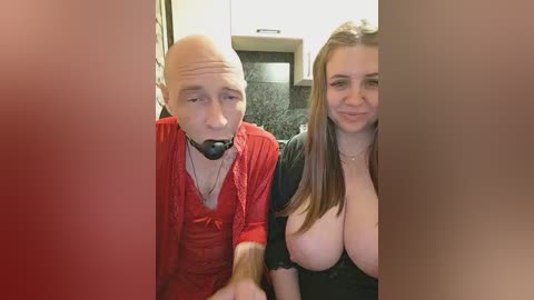 Video of a bald man in a red shirt and black gag, kneeling beside a topless woman with large breasts, in a kitchen setting.
