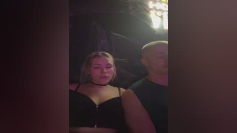 Video of two young people in dimly lit, dark room. A blonde woman with a black choker and black top sits next to a bald man in a green shirt.