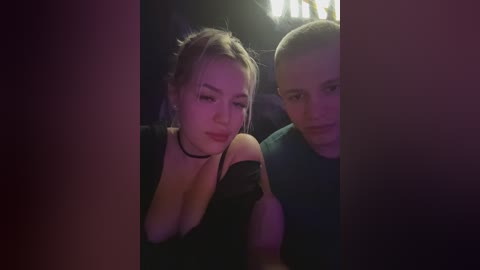 Video of two people in a dimly lit car, showing a woman with blonde hair and a black choker, and a bald man in a blue shirt.