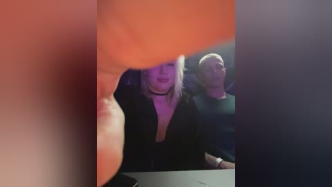 A video shows two people, a woman with blonde hair in a low-cut black dress and a bald man in a dark shirt, seated closely together, partially obscured by a blurred orange object in the foreground.
