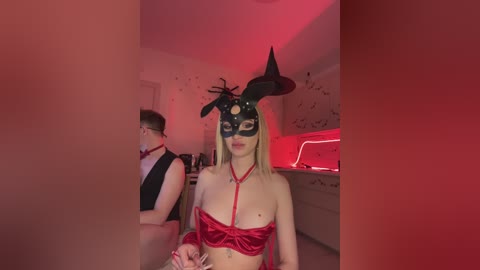 Video of a fair-skinned woman with blonde hair wearing a black, horned mask, red satin lingerie, and a red rope harness. She stands in a dimly lit room with red lighting, a man in a black tuxedo partially visible in the background.