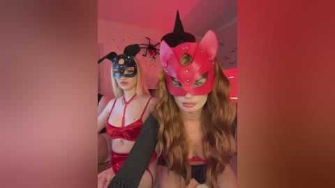 Video of two women in Halloween-themed costumes: one in red, the other in black, both with elaborate masks and lingerie, posing indoors with red lighting.