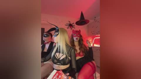 Video of three women in Halloween costumes: one in a black devil mask, another in a black mask, and a third in a red cat mask. They are sitting on a bed with a red glow.