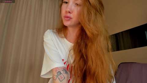 Video of a young woman with long, wavy, reddish-brown hair, wearing a white T-shirt with red text, standing indoors. Her left arm has a tattoo. Background shows beige walls and a window with curtains.
