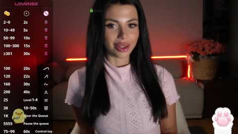 Video of a young woman with straight black hair and a light pink floral dress, captured in a dimly lit living room with a pink vase and red LED strip.