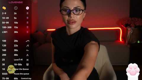 Video of a woman with dark hair, wearing black glasses and a black dress, sitting in a dimly lit room with red neon lights, holding a phone with a \"Loverne\" app.