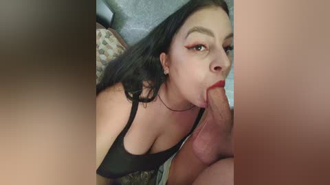 A video shows a young woman with long black hair and a black tank top, performing oral sex on a man with a large penis. The background is blurred, featuring a patterned cushion.