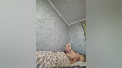 A video of a nude, balding, bearded man with a relaxed posture, lying on a bed with patterned sheets in a small, dimly lit room with textured, light blue walls.