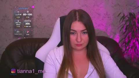 Video of a young woman with long brown hair, wearing a white top, sitting on a black leather couch. Background includes a digital screen displaying health data, a pink lit plant, and a textured wall.