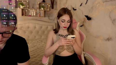Video of a young woman with light skin and straight brown hair, wearing a leopard print bralette and black skirt, sitting on a pink chair, focused on her smartphone, in a cozy, dimly lit room with a beige tufted headboard and a shelf with candles.
