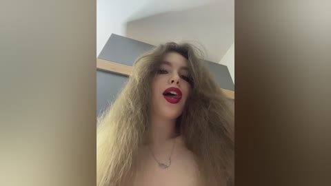 Video of a young woman with voluminous, messy blonde hair, wearing red lipstick, standing indoors against a modern, light-colored background.