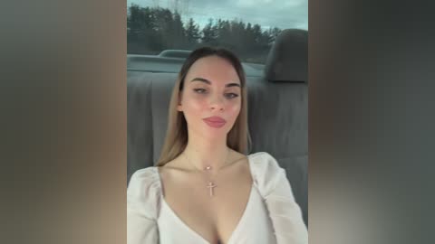 Video of a young woman with light skin, long straight brown hair, wearing a white blouse with a deep V-neck, a cross necklace, and a relaxed expression. Background shows a car interior with a cloudy sky visible through the windows.