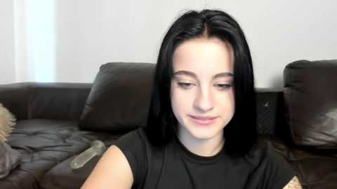Video of a young woman with straight black hair, fair skin, and a light complexion, sitting on a dark leather couch. She wears a black top and has a neutral expression.