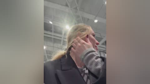 A video captures a blonde woman in a grey coat with a patterned scarf, adjusting her ponytail in a modern, industrial setting with high ceilings and white beams.