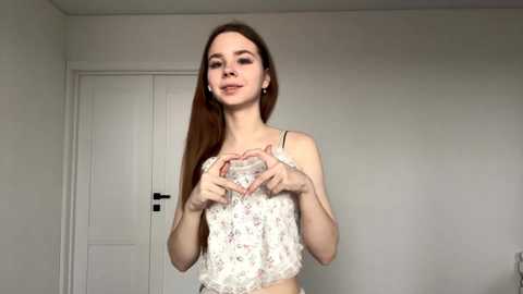 Video of a young Caucasian woman with long brown hair, wearing a floral crop top, making a heart shape with her hands. She stands in a plain, white-walled room with a closed white door in the background.