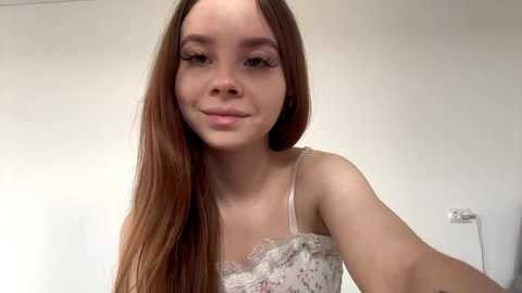 Video of a young woman with long, straight, light brown hair, wearing a white lace camisole, smiling slightly. Background shows a plain white wall and a light switch.